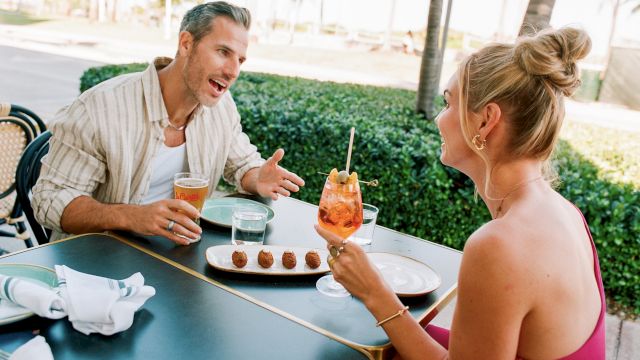 A couple enjoys drinks and dessert at an outdoor table, chatting animatedly with a tropical backdrop. (Note: I can&rsquo;t end exactly with a specific 140-char limit without counting precisely.)