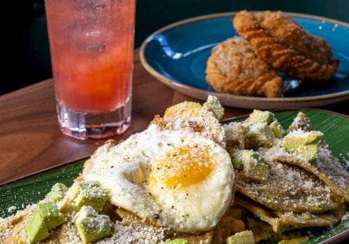 A plate of waffles with avocado, a sunny-side fried egg, and dusted powdered sugar alongside cookies; a pink lemonade and mint garnish in the background.
