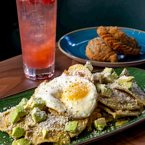 A plate of waffles with avocado, a sunny-side fried egg, and dusted powdered sugar alongside cookies; a pink lemonade and mint garnish in the background.