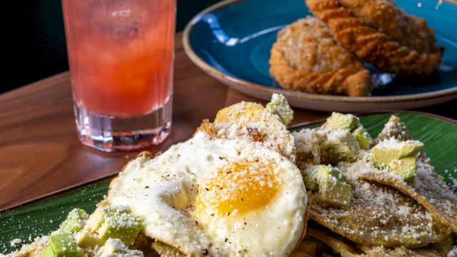 A plate of waffles with avocado, a sunny-side fried egg, and dusted powdered sugar alongside cookies; a pink lemonade and mint garnish in the background.
