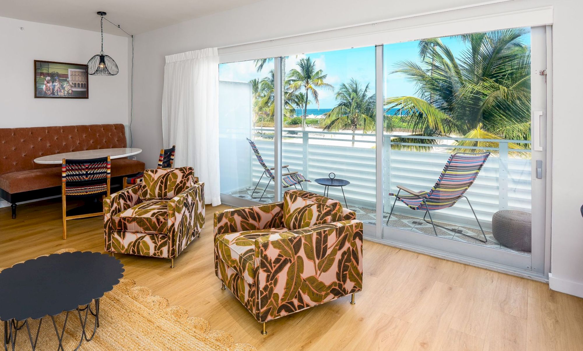 A bright mid-century living room with patterned armchairs, a round black coffee table, a brown sofa, a floor lamp, and a large sliding door opening to a balcony with palm trees outside.