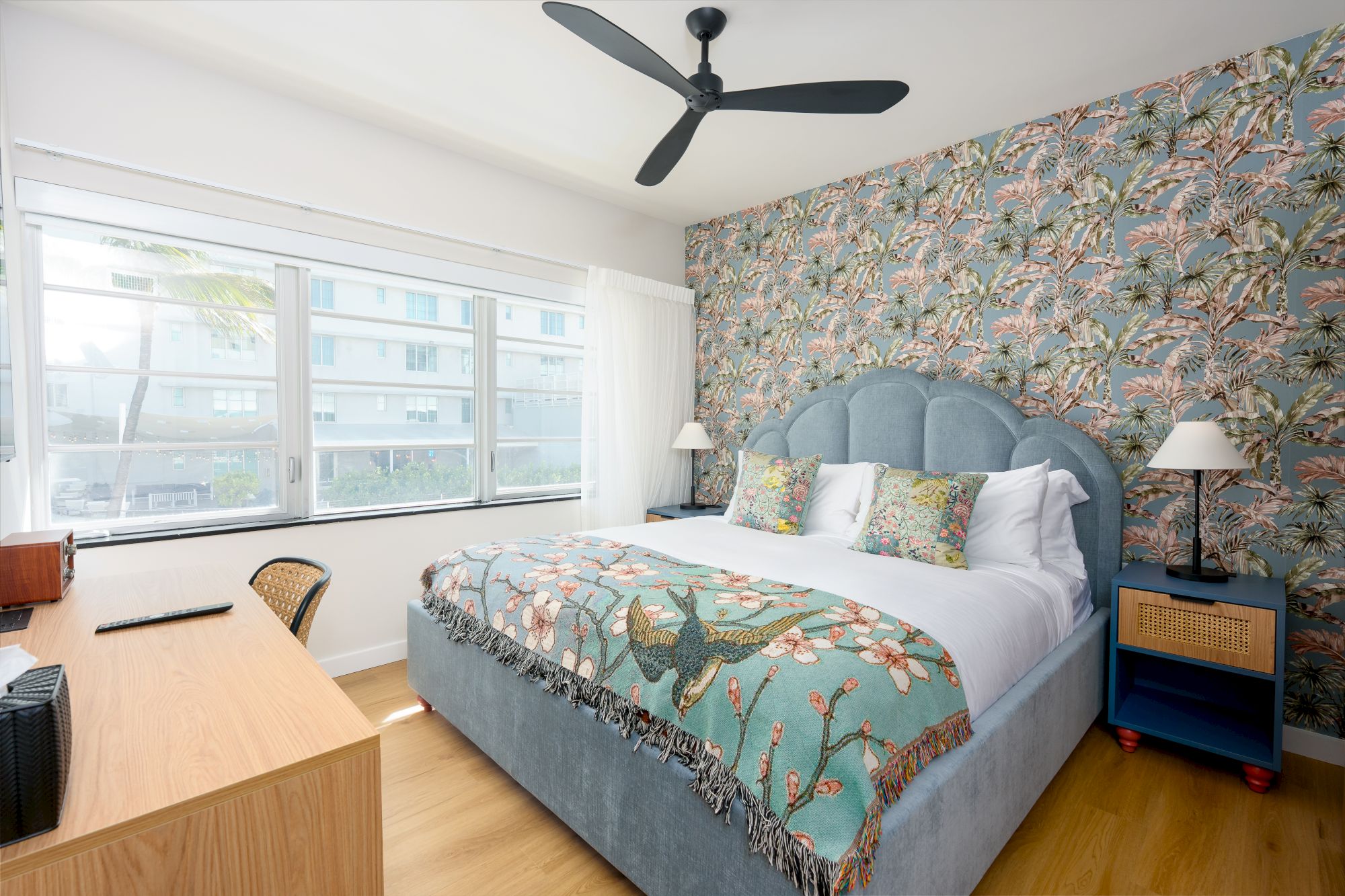 A bright hotel-style bedroom with a blue upholstered bed, floral wallpaper accent wall, large window, ceiling fan, and matching bedside tables with lamps.