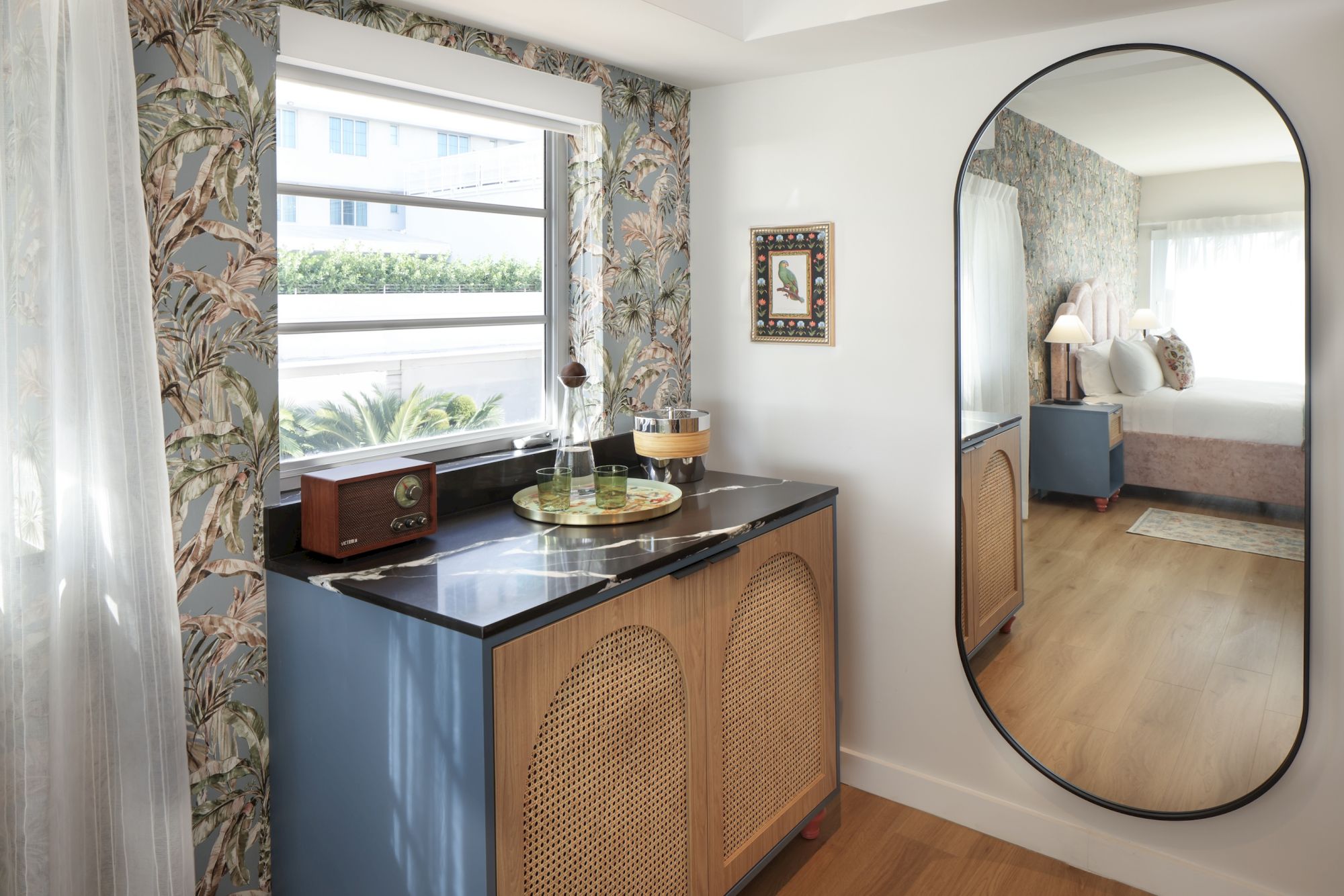 A cozy corner with a blue cabinet, woven rattan doors, a black countertop, a window with floral curtains, a small radio, and a decorative mirror reflecting a bed.