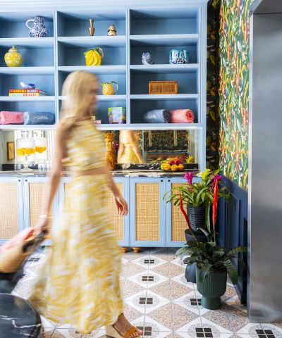Top it at 140 characters, ending with a period: A blurred woman in a yellow dress walks past blue open shelves and a patterned tropical kitchen, plants nearby.