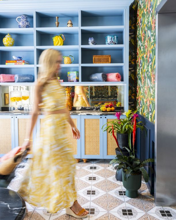 Top it at 140 characters, ending with a period: A blurred woman in a yellow dress walks past blue open shelves and a patterned tropical kitchen, plants nearby.