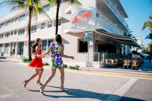 Two women in colorful dresses walk across a sunny street in front of a modern building with palm trees, a mural, and outdoor seating.