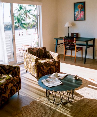 A sunlit living room with patterned armchairs, a green coffee table, a desk and chair, wall art, and sliding doors opening to a sunny outdoor scene.