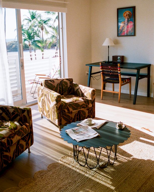 A sunlit living room with patterned armchairs, a green coffee table, a desk and chair, wall art, and sliding doors opening to a sunny outdoor scene.