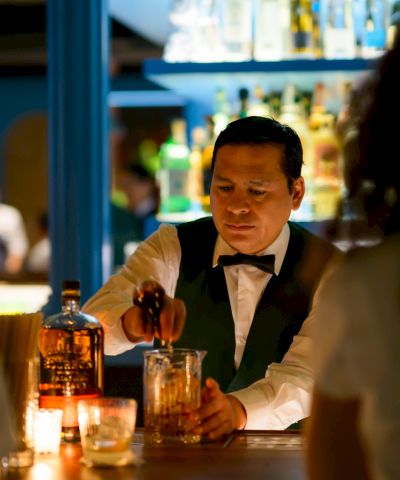 A bartender in a suit pours a drink at a dimly lit bar, candlelit table, patrons in the background, warm and intimate ambiance.
