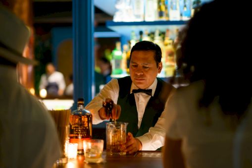 A bartender in a suit pours a drink at a dimly lit bar, candlelit table, patrons in the background, warm and intimate ambiance.