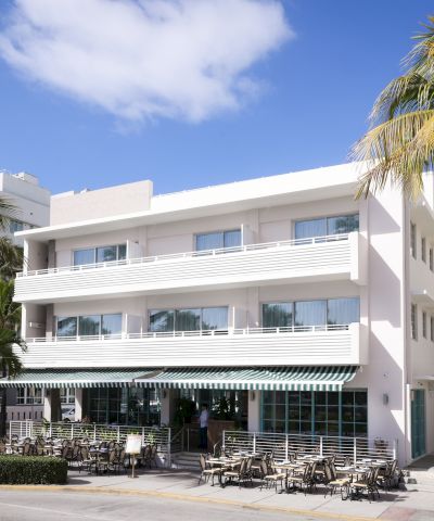 A sunny beachfront hotel with white balconies, palm trees, and outdoor seating ready for guests. Top it at 140 characters, always ending the sentence.