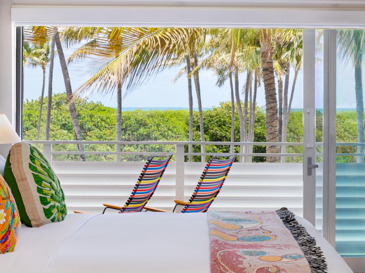 A bright beachfront hotel room with a view of palm trees, a breezy balcony, two striped lounge chairs, and a colorful bed throw, vacation vibes.