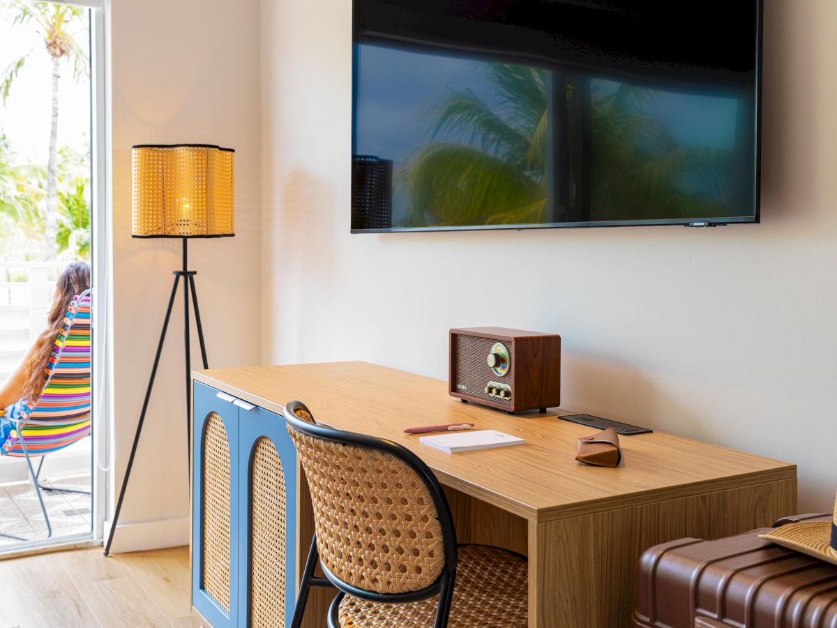 A modern hotel room with a desk, two chairs, a wall-mounted TV, vintage radio, and a floor lamp; glass door opens to a sunny outdoor area.