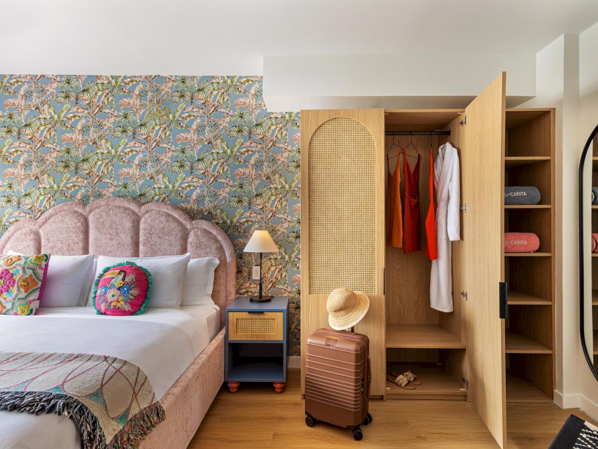 A cozy hotel-style bedroom with a floral accent wall, a pink tufted bed, colorful pillows, a wooden wardrobe, and a small suitcase on the floor.