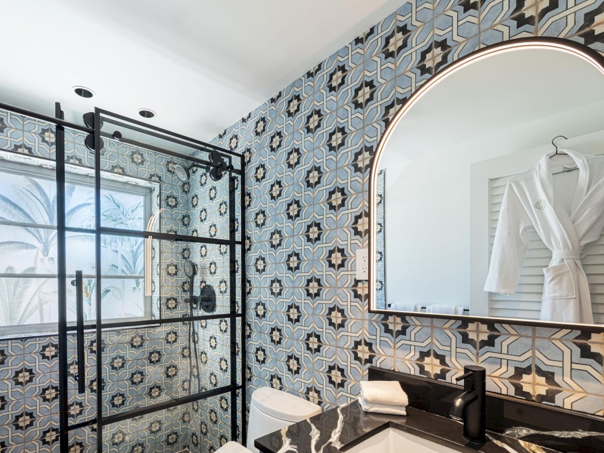 A stylish bathroom with bold geometric patterned tiles, a glass shower enclosure, a black countertop, and a rounded mirror above a white robe hanging on the door, all under bright light.