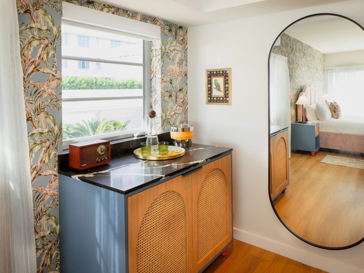 A cozy corner with a blue cabinet, woven doors, a monitor/window view, plant, vintage radio, lamp, and a large oval mirror reflecting a bedroom.
