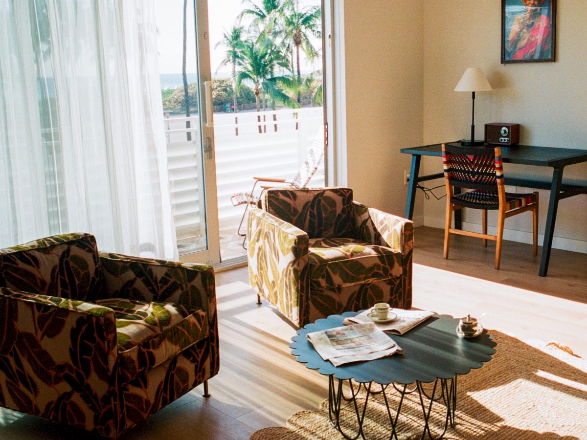 A bright living room with two patterned armchairs, a round coffee table, a desk and chair near a window, and a sliding glass door to a balcony.