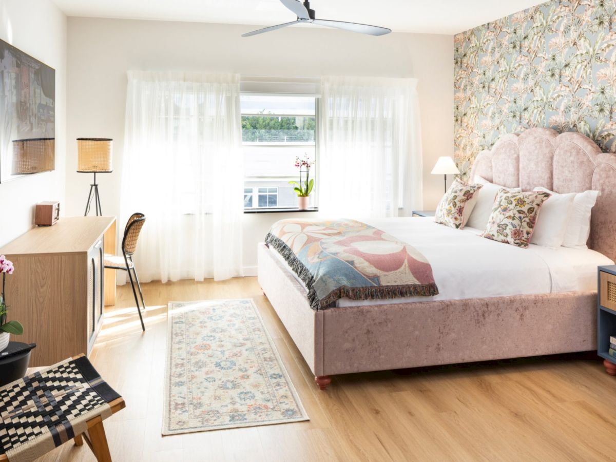 A bright, stylish hotel-style bedroom with a pink upholstered bed, floral wallpaper accent wall, wooden floors, two nightstands, a desk and chair, a patterned rug, and airy sheer curtains by a window.