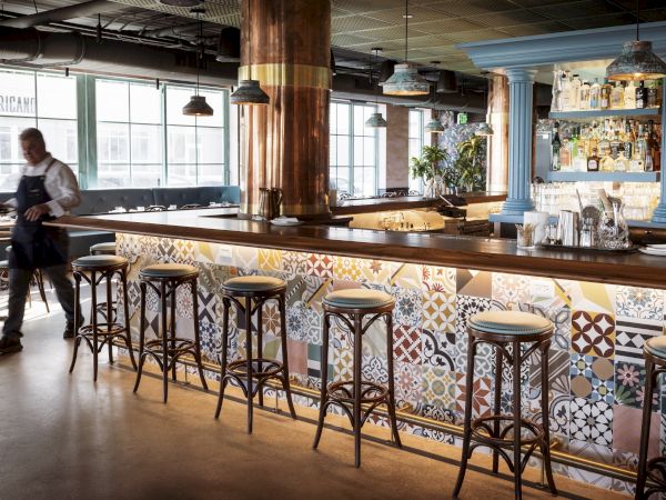 A cozy bar with patterned tile counter, wooden stools, blue accents, and large windows; a waiter adjusts the scene in a calm, airy space.