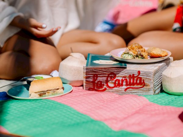A picnic scene with snacks on plates and a stack of cards labeled &ldquo;La Cantina,&rdquo; set on a colorful blanket with people lounging nearby.