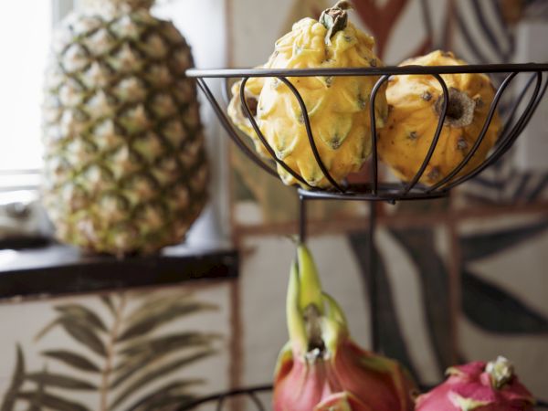 A pineapple sits on the left, while a two-tier fruit stand holds yellow limes/pears on top and pink dragon fruit on the bottom, against leafy wallpaper.