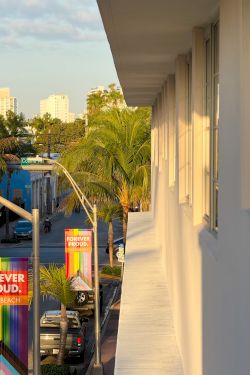 A sunny street view from a balcony, with palm trees, colorful rainbow Pride banners, parked cars, and buildings in the distance.