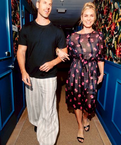 Couple walking down a hallway: man in black shirt with white striped pants, woman in black floral dress, both smiling as they stroll. end sentence.