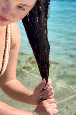 A girl with sun-kissed freckles smiles at the camera while gripping long wet hair on a sunny beach with turquoise water behind her, enjoying the moment.
