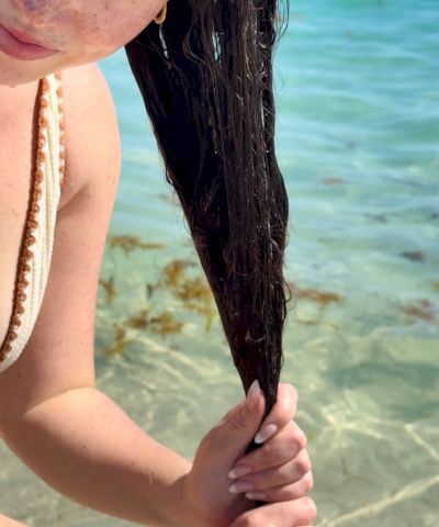 A girl with sun-kissed freckles smiles at the camera while gripping long wet hair on a sunny beach with turquoise water behind her, enjoying the moment.
