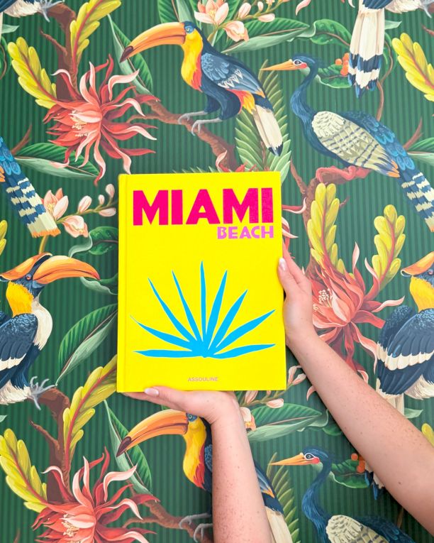 Colorful tropical wallpaper with parrots and flowers, holding a bright yellow Miami Beach booklet.