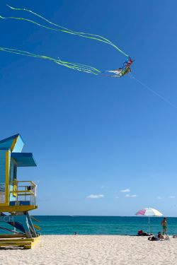 Kite surfer soaring above the beach with long green and blue tails, a lifeguard stand on the left, and people relaxing near the shore.