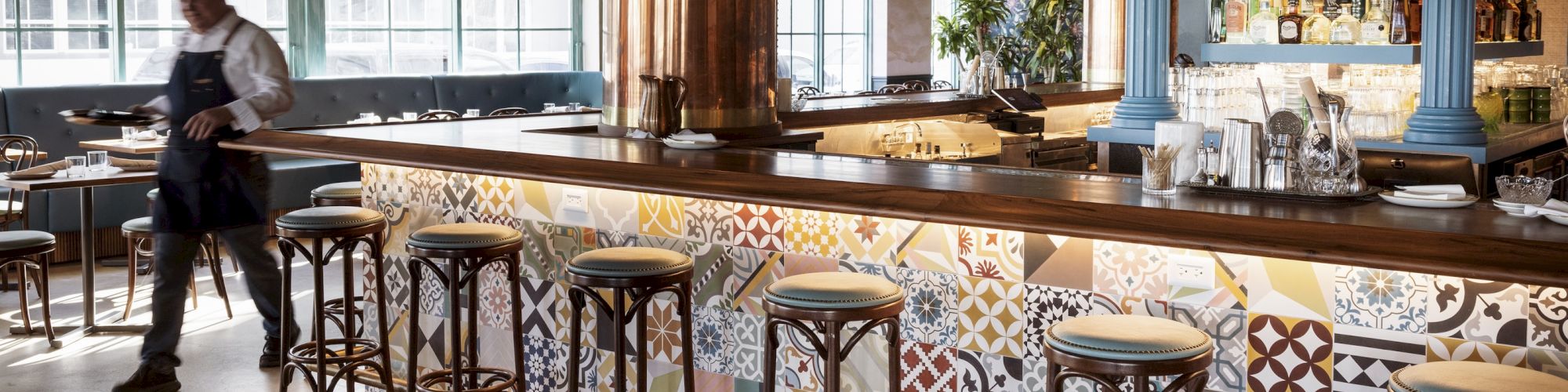 A stylish empty bar with mosaic tiled counter, tall stools, warm lighting, and a bartender near the left window, ready to serve.