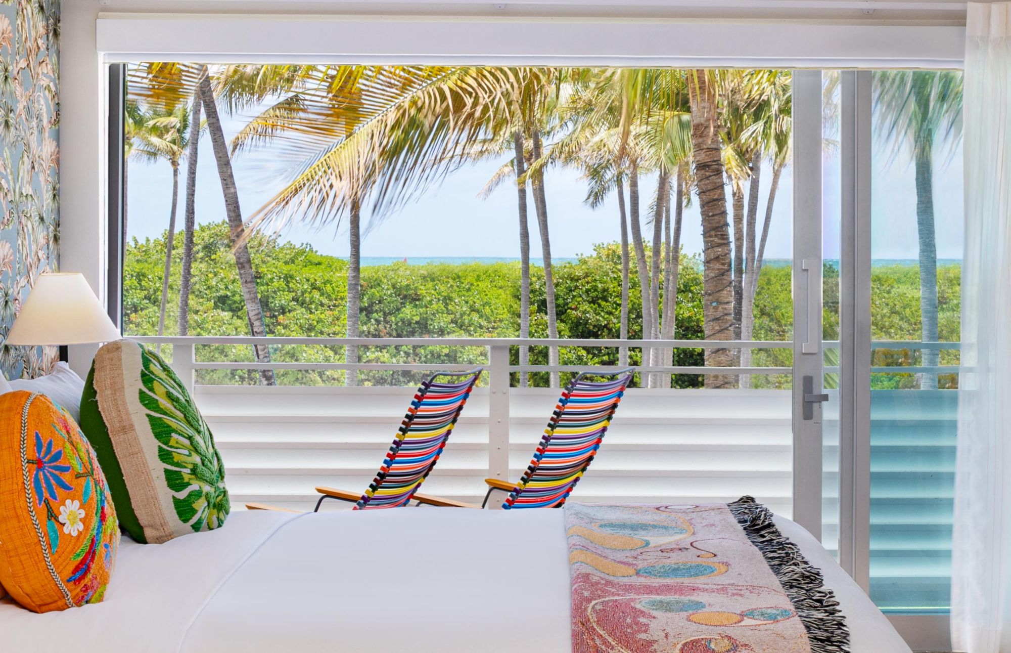 A bright bedroom overlooks a tropical view with palm trees, a balcony, and two colorful lounge chairs by the sea, creating a sunny retreat.