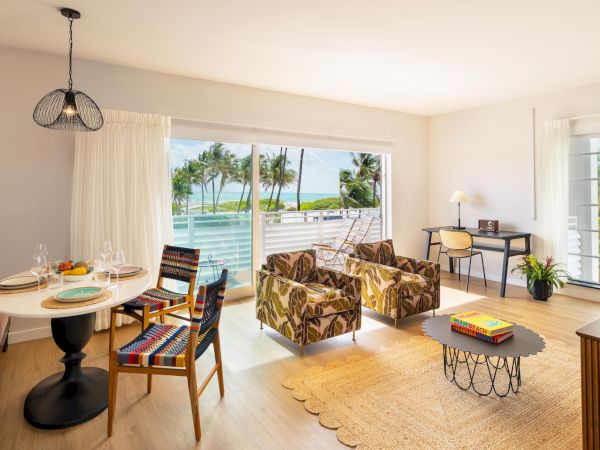 A bright, airy living-dining room with a large sliding door to a beach view, tropical chairs, a round dining table, and a cozy seating area.