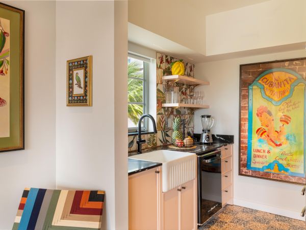 A compact kitchen with a farmhouse sink, open shelves, and a colorful vintage poster on the wall, plus a striped chair and framed art.
