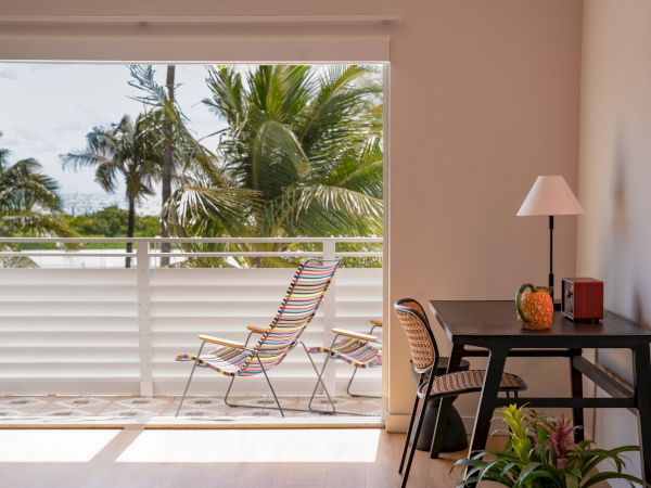 A bright balcony room with a desk and chair, potted plant, and a view of palm trees on a sunny day; two chairs sit on the patio outside.