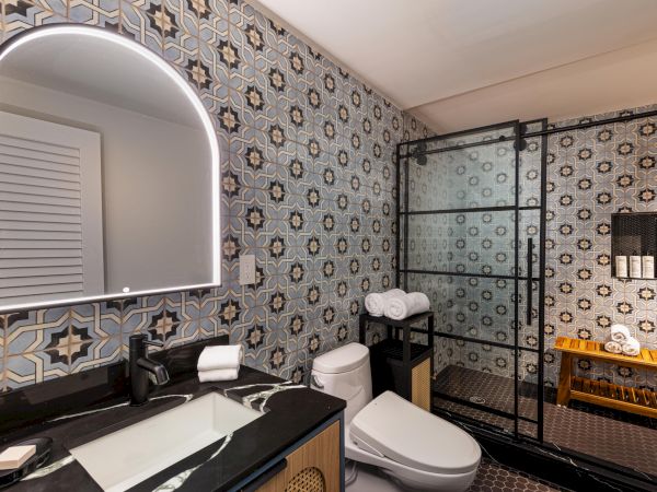 A stylish bathroom with patterned tile walls, a black dual-sink vanity, round backlit mirror, a modern toilet, and a glass-walled shower area, all neatly arranged.