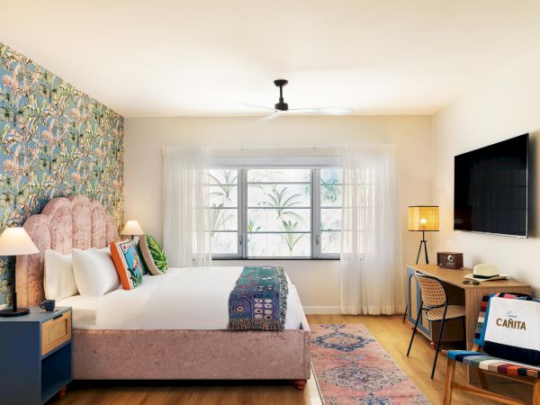 A cozy hotel-style bedroom with a floral accent wall, plush pink upholstered bed, bright window, desk with chair, and mounted TV, airy and inviting.