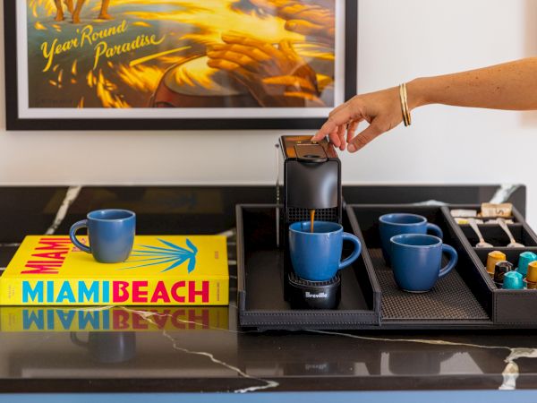 A person prepares coffee with a Nespresso machine; blue mugs and a yellow Miami Beach book sit beside a tray of pods.