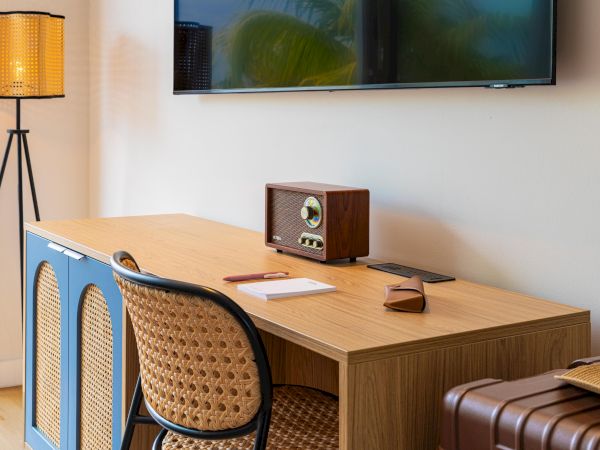 A mid-century desk setup with a wooden table, blue cabinet panels, a woven chair, a retro radio, a notebook, a pair of glasses, a tall tripod lamp, and a wall-mounted TV above.