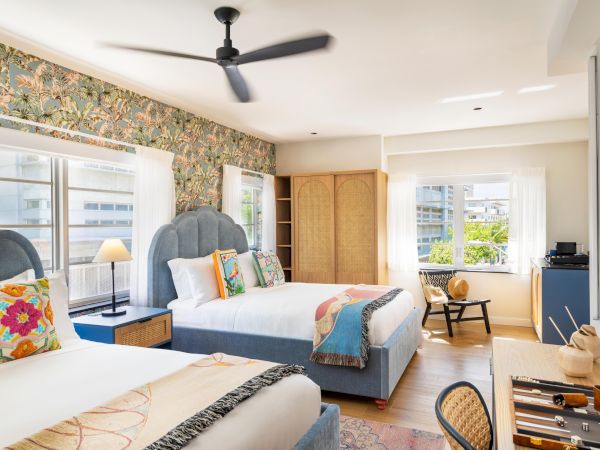 A bright hotel room with two queen beds, patterned headboards, colorful throw pillows, a ceiling fan, wooden furniture, a desk, window seats, and sunny view outside.