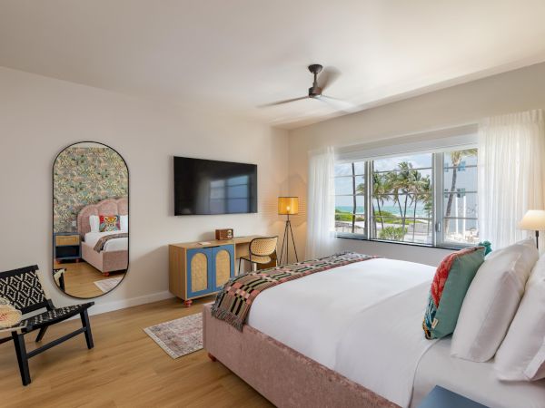 A bright hotel room with a king bed, flat-screen TV, a seating chair, a dresser, a large window view of palm trees, and a ceiling fan.