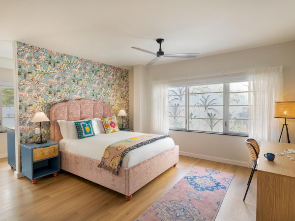 A bright, stylish hotel-style bedroom with a pink upholstered bed, floral wallpaper accent wall, blue nightstands, desk, and large windows letting in natural light, ending with a cozy, airy vibe.