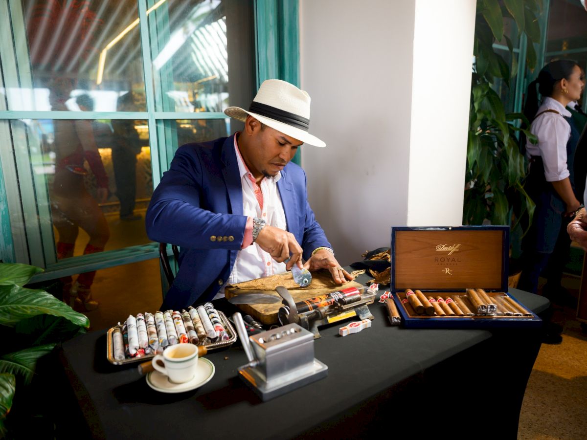 A man in a blue blazer and hat arranges cigars on a table with a portable display, coffee cup, and a brown case nearby.