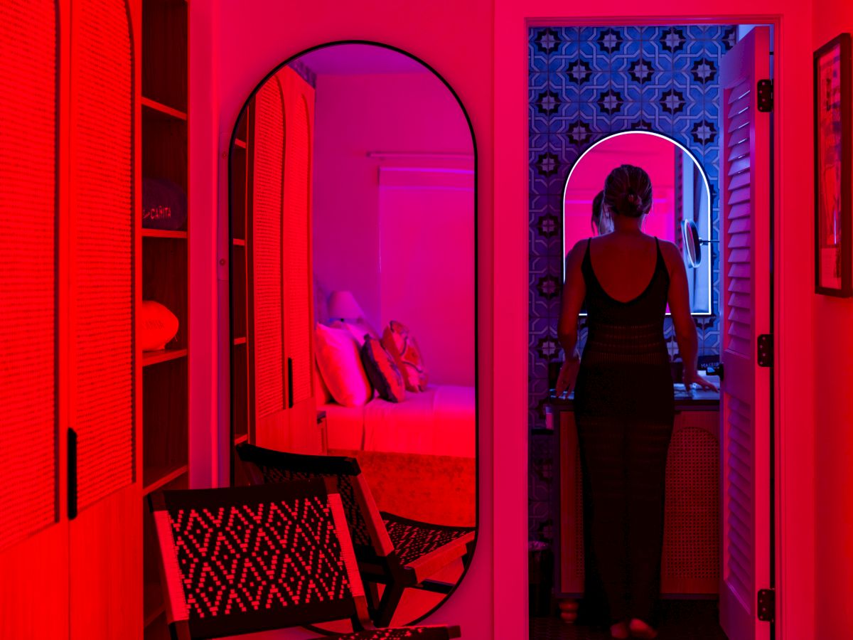 A woman in a black outfit stands in a doorway, bathed in red and purple neon lights reflecting across a stylish, mirrored interior.