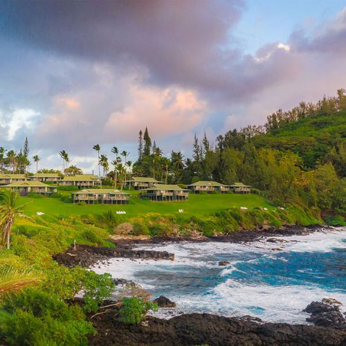 A tropical seaside with rocky shore, lush green hillside, and white buildings perched above the water at sunset.