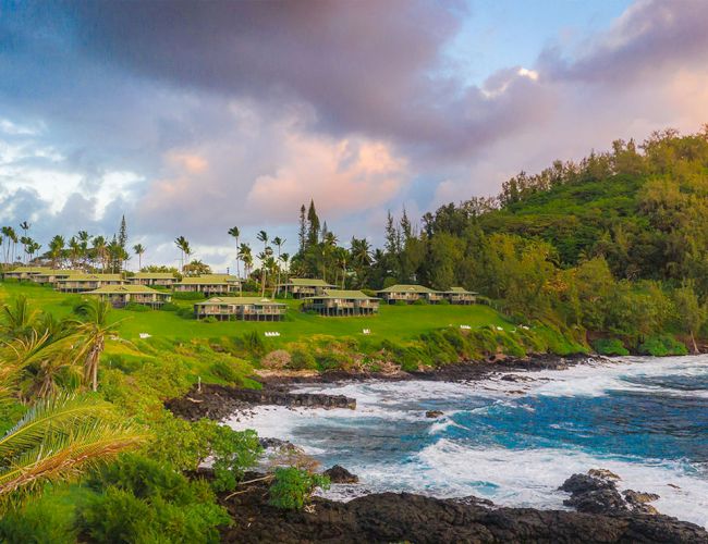 A tropical seaside with rocky shore, lush green hillside, and white buildings perched above the water at sunset.