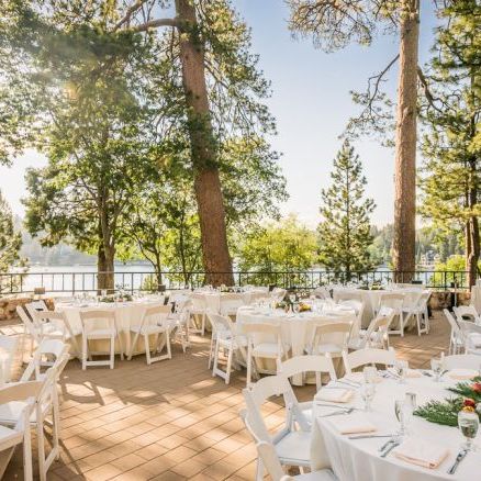 An outdoor dining setup with many round tables, white chairs, and elegant table settings under tall trees by a sunny lakeside.