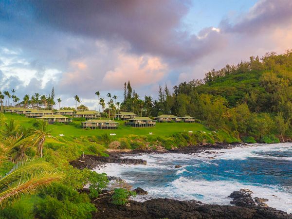 Coastal resort on a rocky shoreline with green hills, palm trees, and low-rise houses overlooking blue waves under a partly cloudy sky.