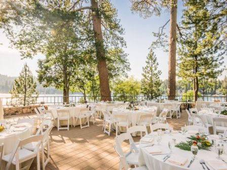 An elegant outdoor event setup with white-draped tables, chairs, and floral centerpieces on a sunlit terrace amid trees overlooking water.